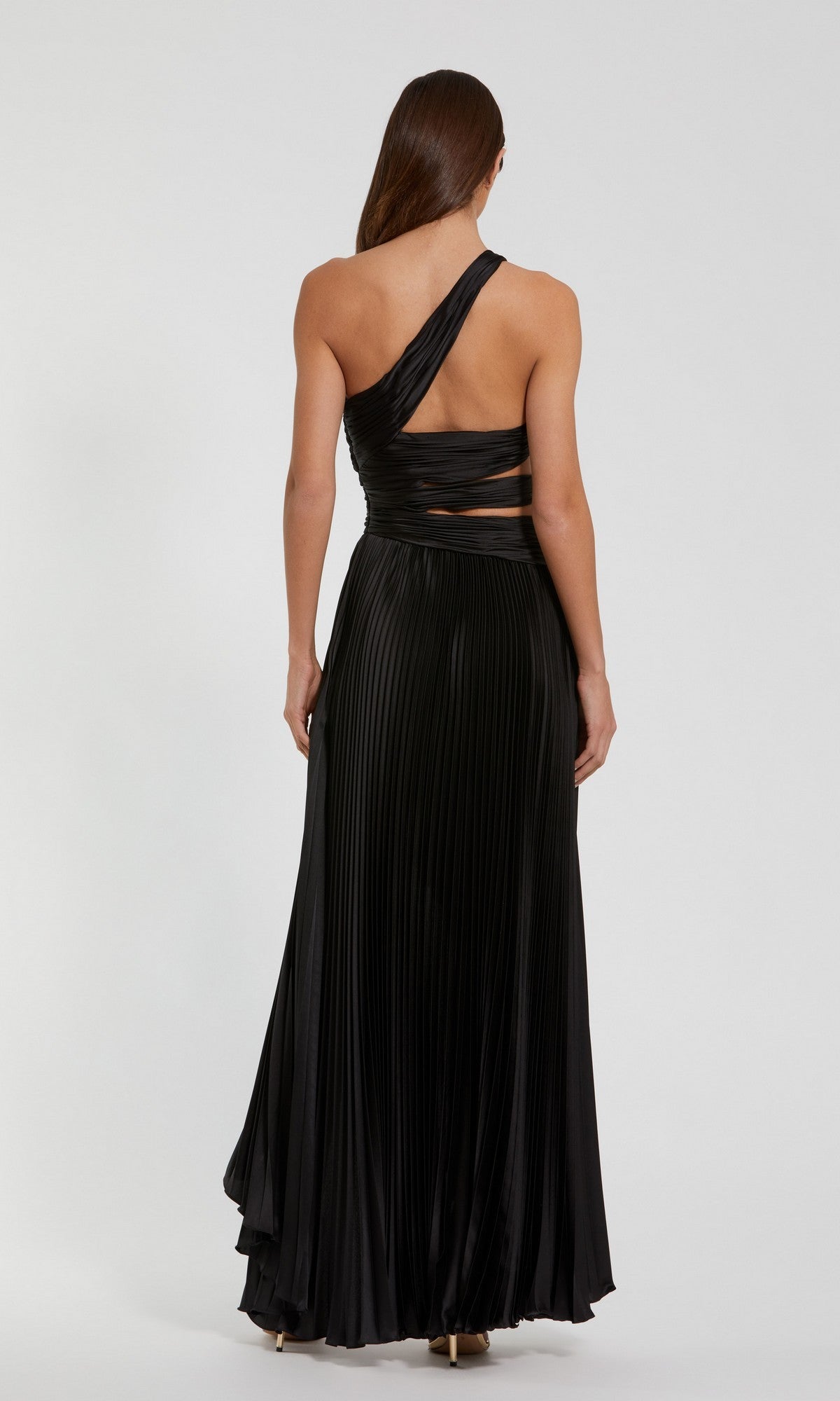 A woman stands facing away, wearing the Mac Duggal 49837 High-Low Pleated Formal Dress with cut-out and waist details, set against a plain light background.